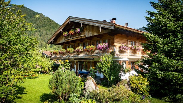 A wooden chalet with a balcony, surrounded by lush greenery and a mountain backdrop.