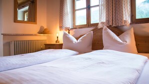 A double bed with white linens and two pillows, a bedside table with a lamp, a mirror on the wall, and a window with curtains.