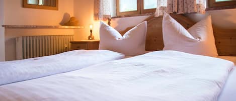 A double bed with white linens and two pillows, a bedside table with a lamp, a mirror on the wall, and a window with curtains.