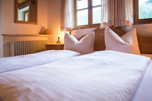 Een tweepersoonsbed met witte lakens en twee kussens, een nachtkastje met een lamp, een spiegel aan de muur en een raam met gordijnen.