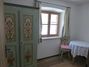 A room with a window, a door with floral designs, a small table with a tablecloth, and a chair. - Vacation apartment 3 Sepp - Guesthouse Becher, Kreuth-Point (Kreuth)