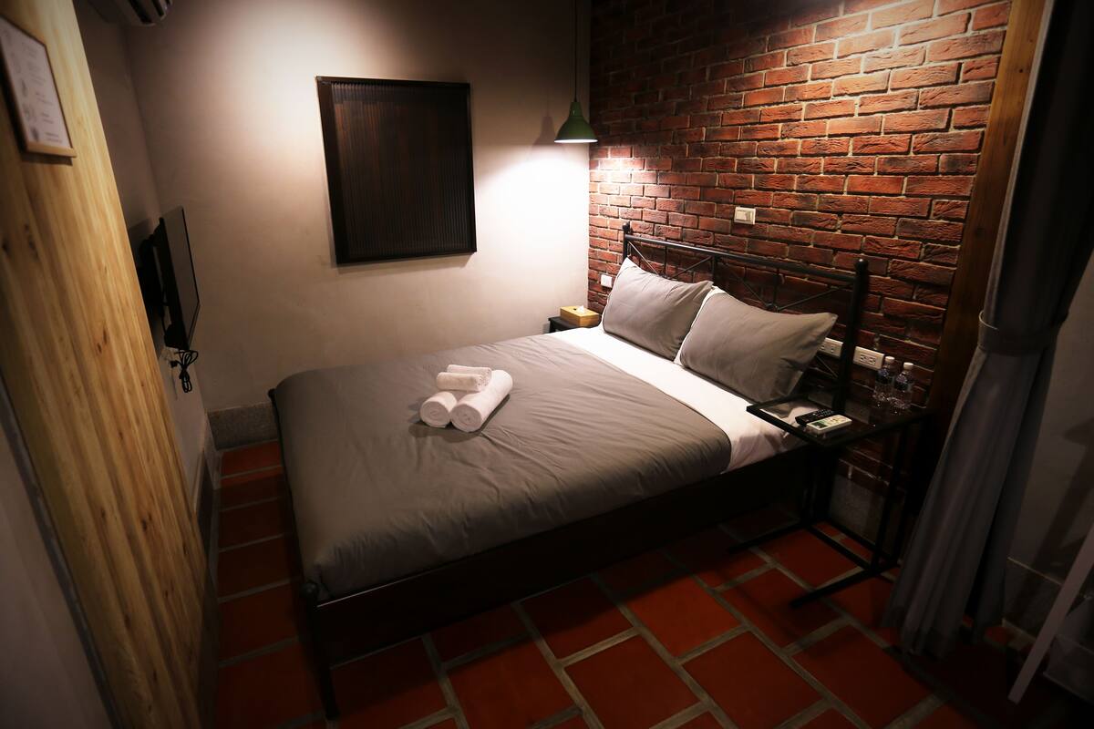 A neatly made bed with pillows and towels, a brick wall, a wooden door, and a nightstand with a lamp.
