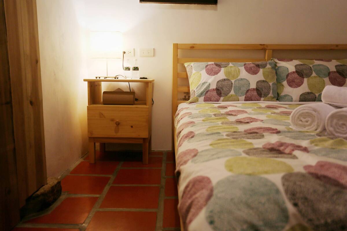 A bedroom with a wooden bed, a nightstand with a lamp, and a small wooden drawer unit.
