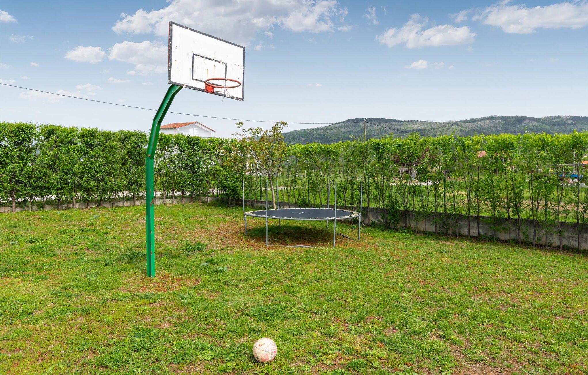 Ein Basketballkorb, eine Hüpfburg und ein Fußball in einer grünen Freifläche mit Bäumen und Hügeln im Hintergrund.