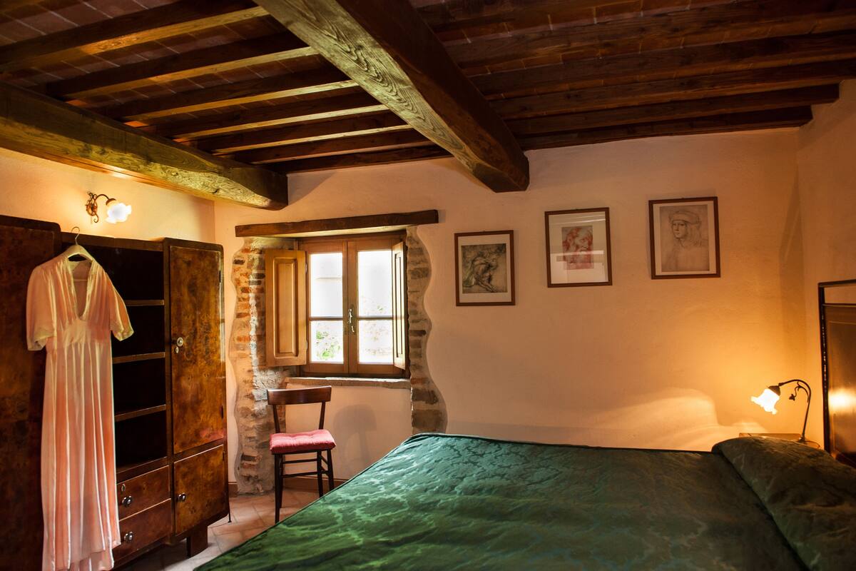 A bedroom with a bed, a wooden wardrobe, a chair, a window, and a wall-mounted light fixture.
