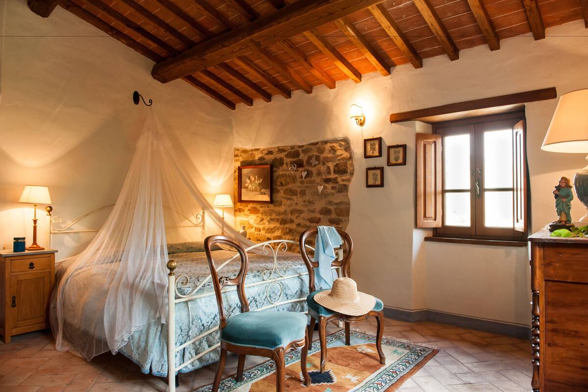 A bedroom with a bed under a canopy, a nightstand, a chair with a hat, a dresser, and a window with shutters.