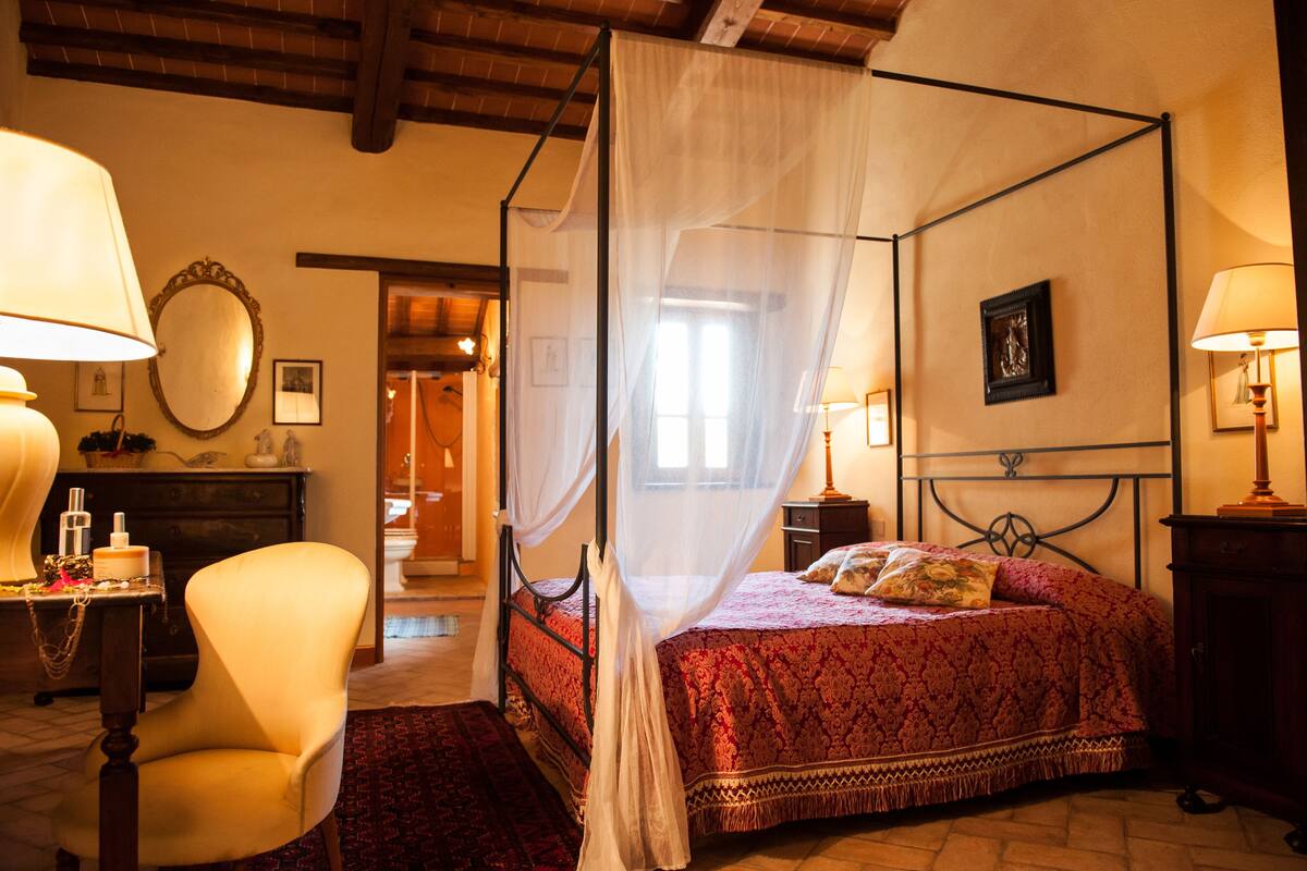 A four-poster bed with a canopy, a chair, a nightstand, and a lamp.