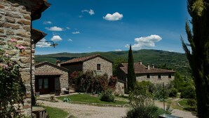 A stone building with a gravel driveway, surrounded by greenery and hills in the background. - Agriturismo Azienda Agricola il Pozzo (Capolona)