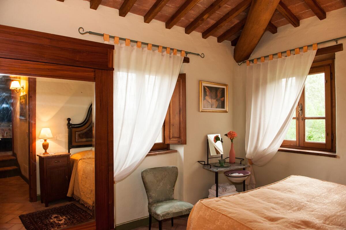 A bedroom with a bed, a chair, a bedside table with a lamp, a mirror, and curtains.