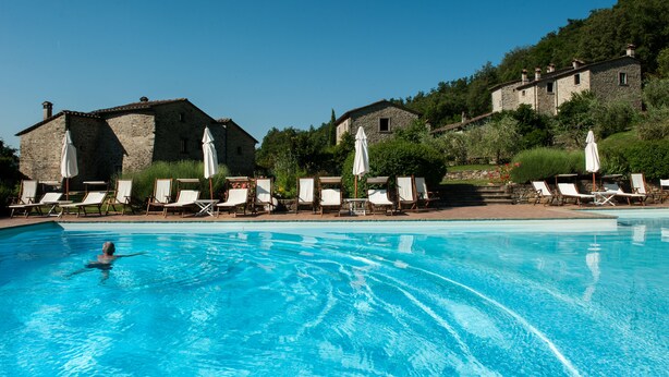 A swimming pool with lounge chairs and umbrellas, surrounded by stone buildings and greenery. - Agriturismo Azienda Agricola il Pozzo (Capolona)