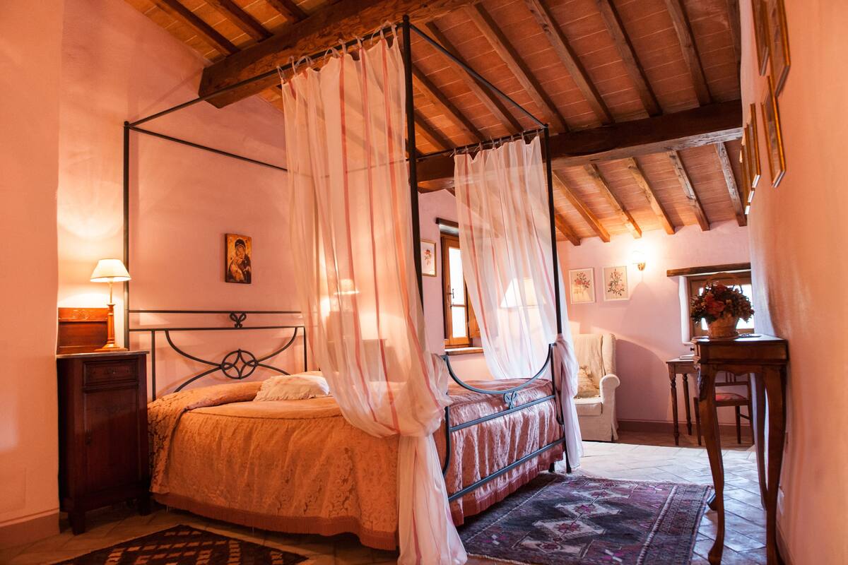 A bedroom with a four-poster bed, a nightstand, a chair, and a table with a vase of flowers.