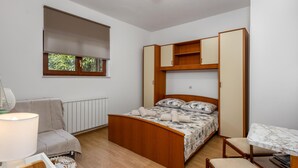 Quarto compacto com cama, secretária pequena, cadeira, radiador e janela com vista para vegetação.