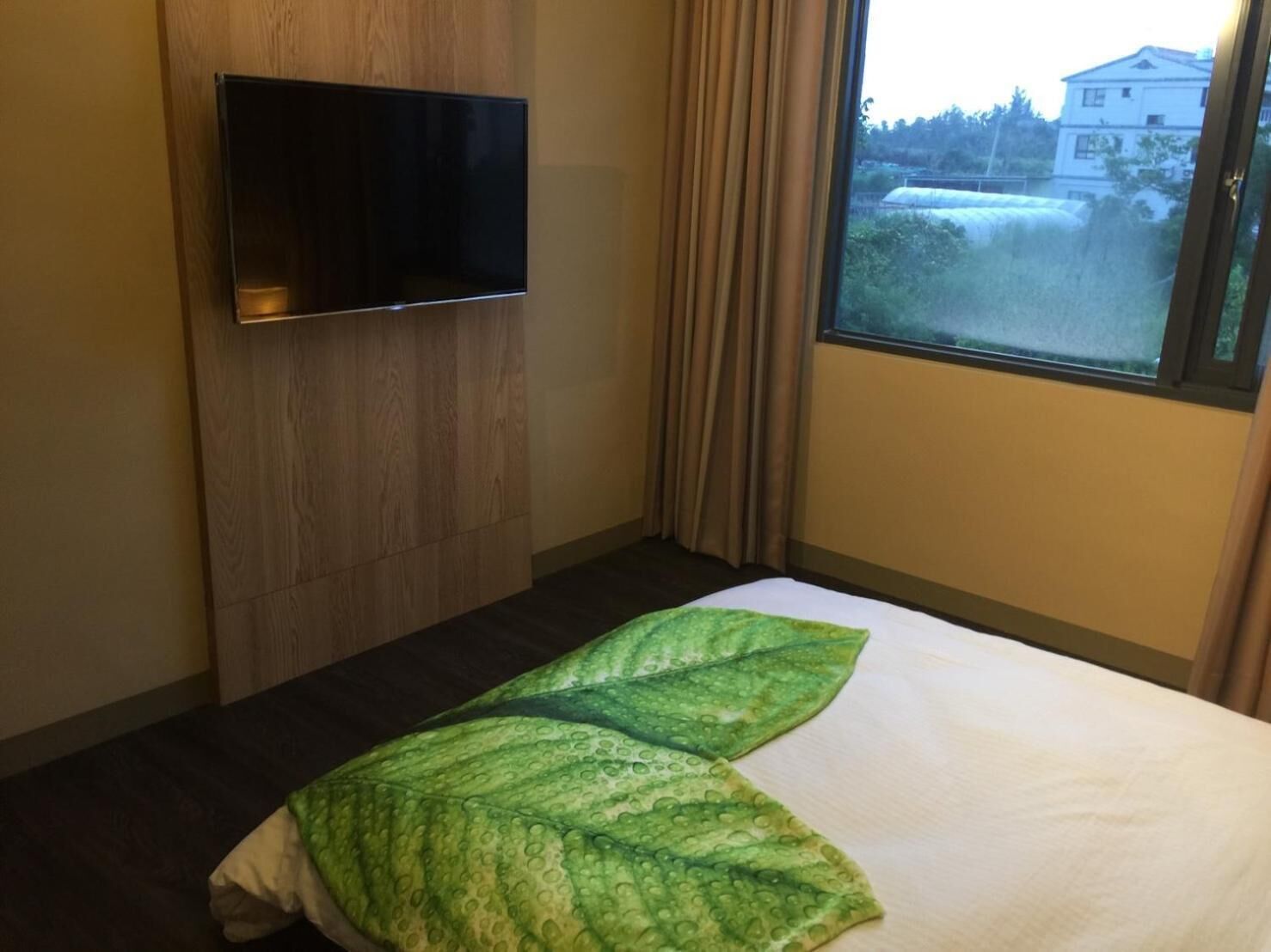 Een hotelkamer met een bed, een flatscreen televisie bevestigd aan een houten paneel, en een raam met gordijnen.