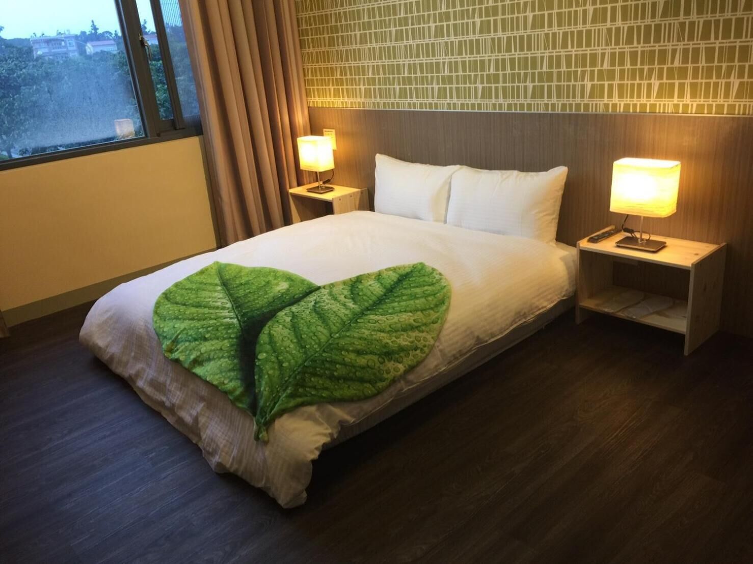 A hotel room with a large bed, two bedside lamps, a green leaf-shaped throw pillow, and a view of trees through the window.