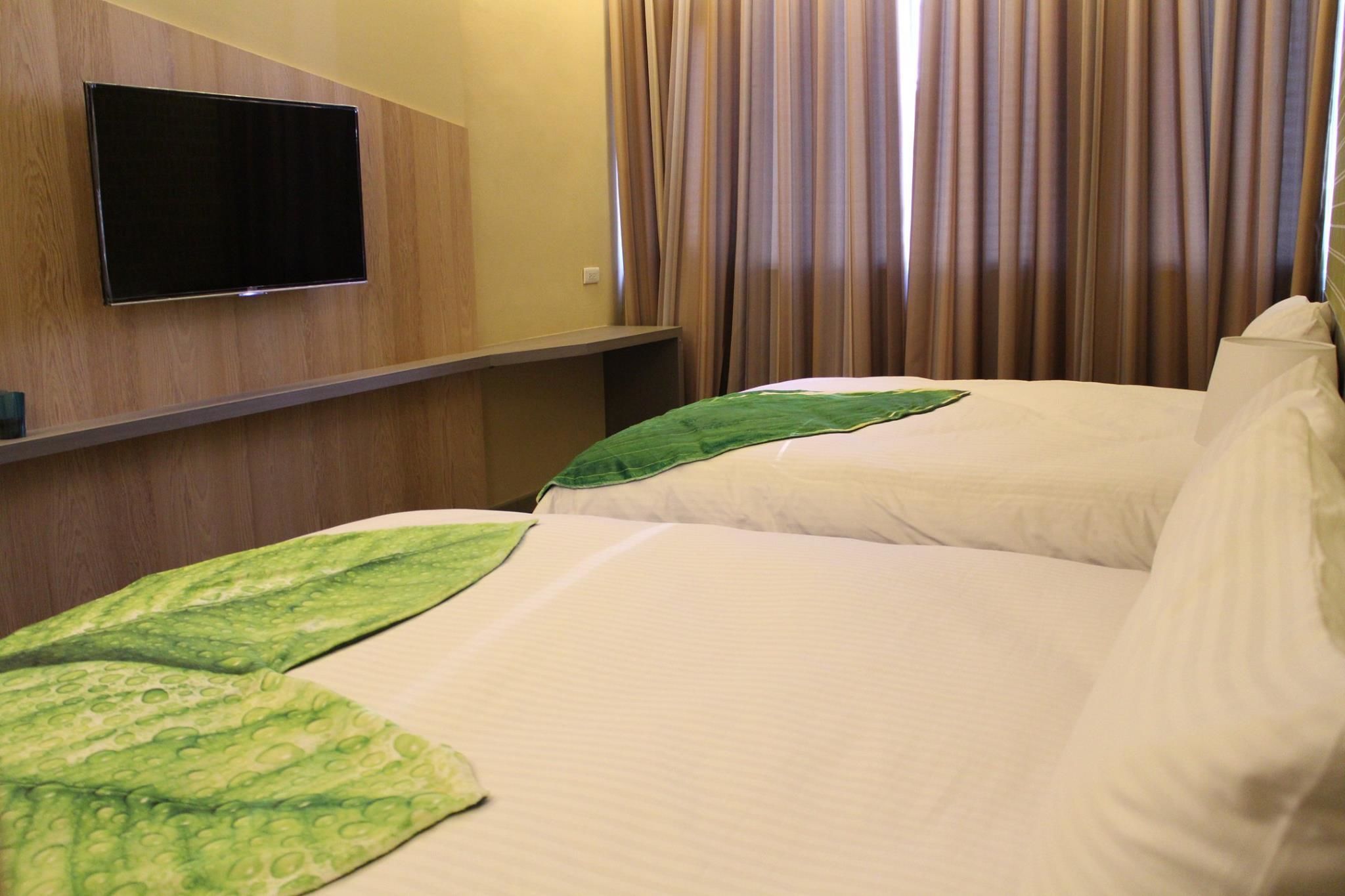 A hotel room with a flat-screen TV mounted on the wall, a bed with white bedding and a green leaf-patterned throw, and a window with vertical blinds.