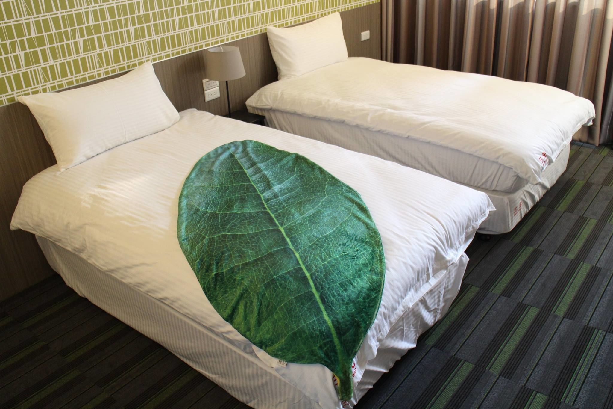 A hotel room with two single beds, a green leaf-shaped pillow, a striped carpet, and a green and white tiled headboard.