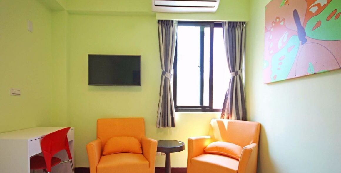A room with two orange chairs, a small round table, a TV mounted on the wall, and a window with curtains.