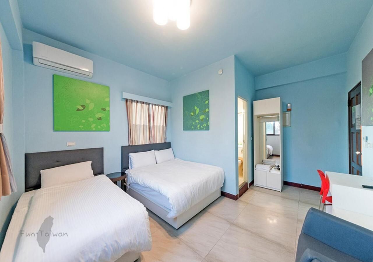 Quarto de hotel com duas camas, uma mesa e uma cadeira. Há uma janela com cortinas, um aparelho de ar condicionado e obras de arte nas paredes.