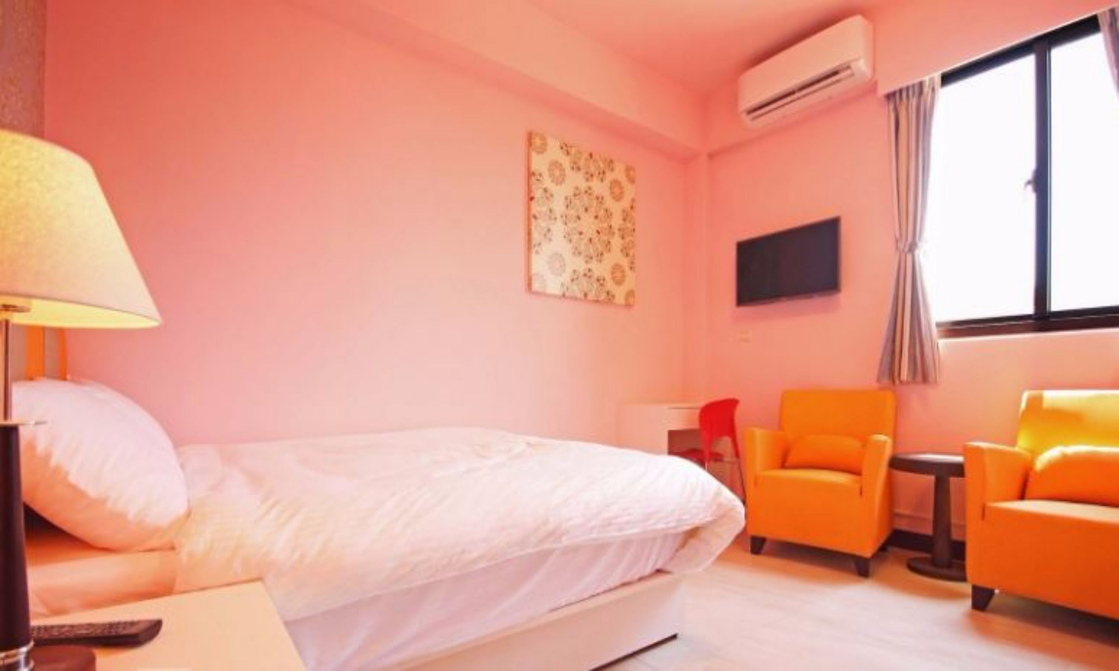 A hotel room with a bed, an orange armchair, a red chair, a small table, and an air conditioning unit.