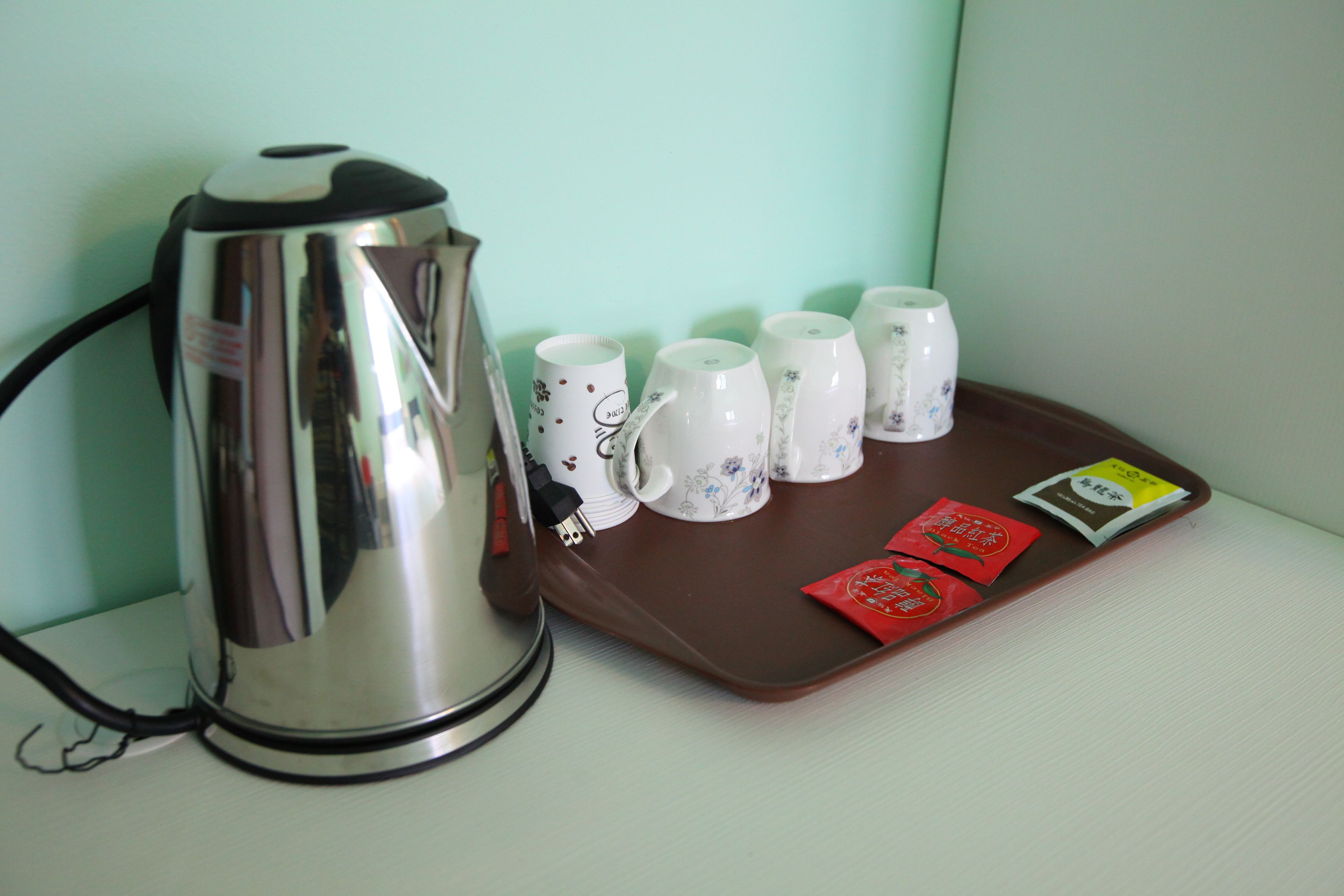 Una tetera eléctrica de acero inoxidable, seis tazas con estampado floral y dos sobres de té sobre una bandeja marrón.