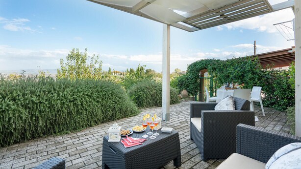 Een terras buiten met wicker meubilair, een tafel gedekt met eten en drinken, en uitzicht op het landschap.