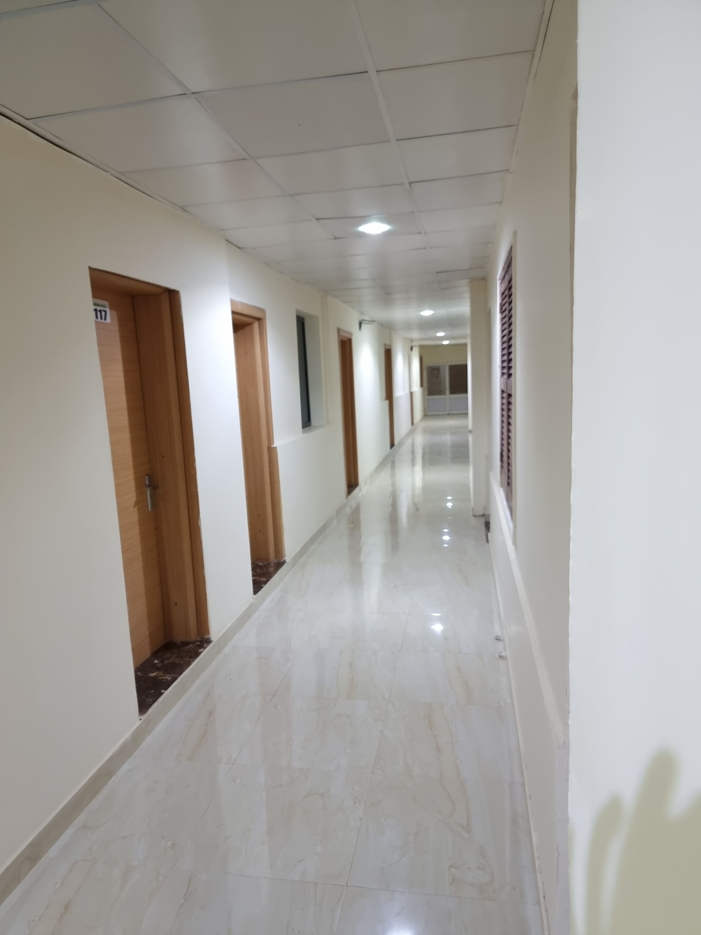 Un pasillo largo y estrecho con paredes blancas y suelo de baldosas brillantes. Hay varias puertas a lo largo del pasillo, algunas con ventanas en la parte superior.