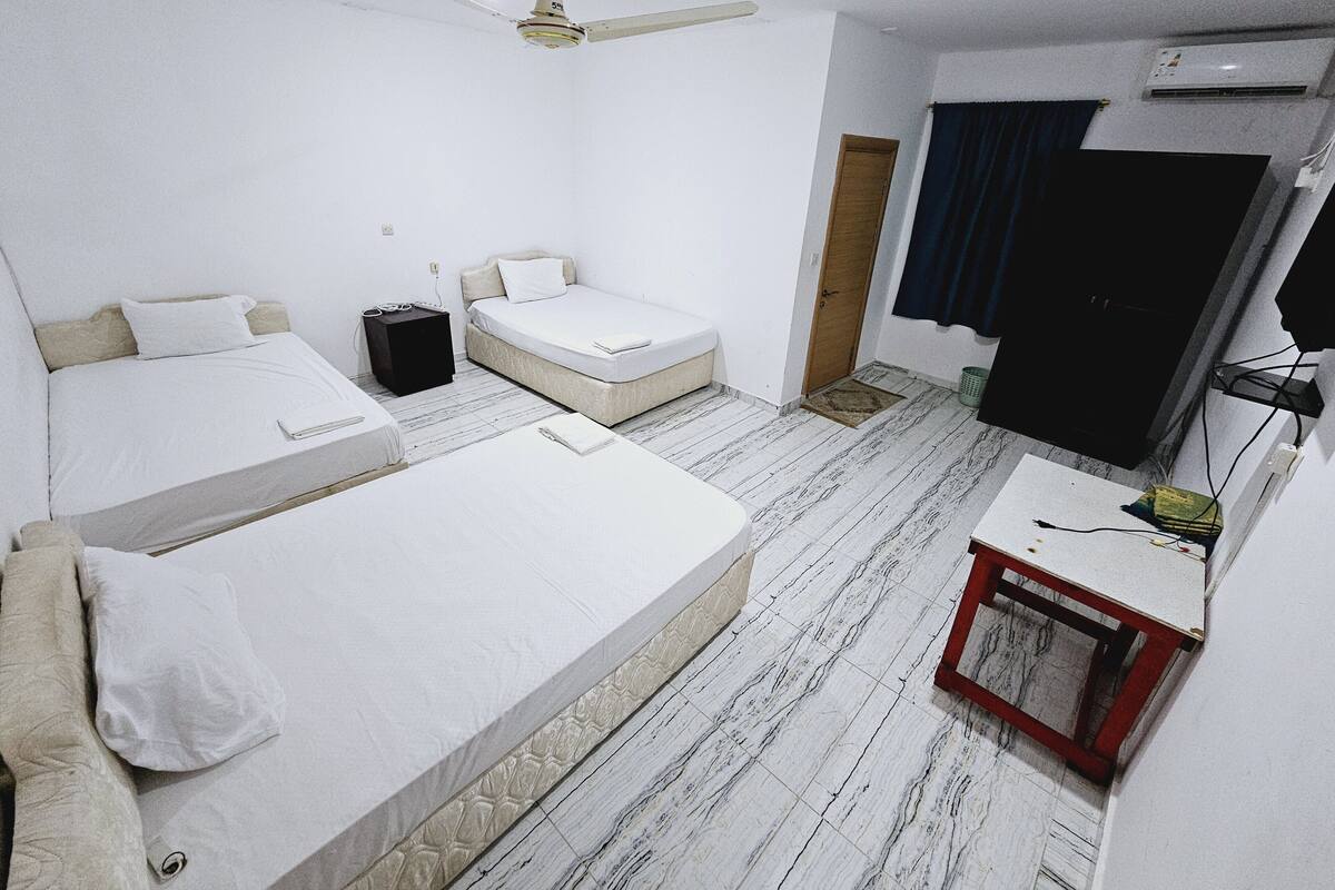 A hotel room with two single beds, a small desk with a chair, a ceiling fan, and a door leading to a dark room.