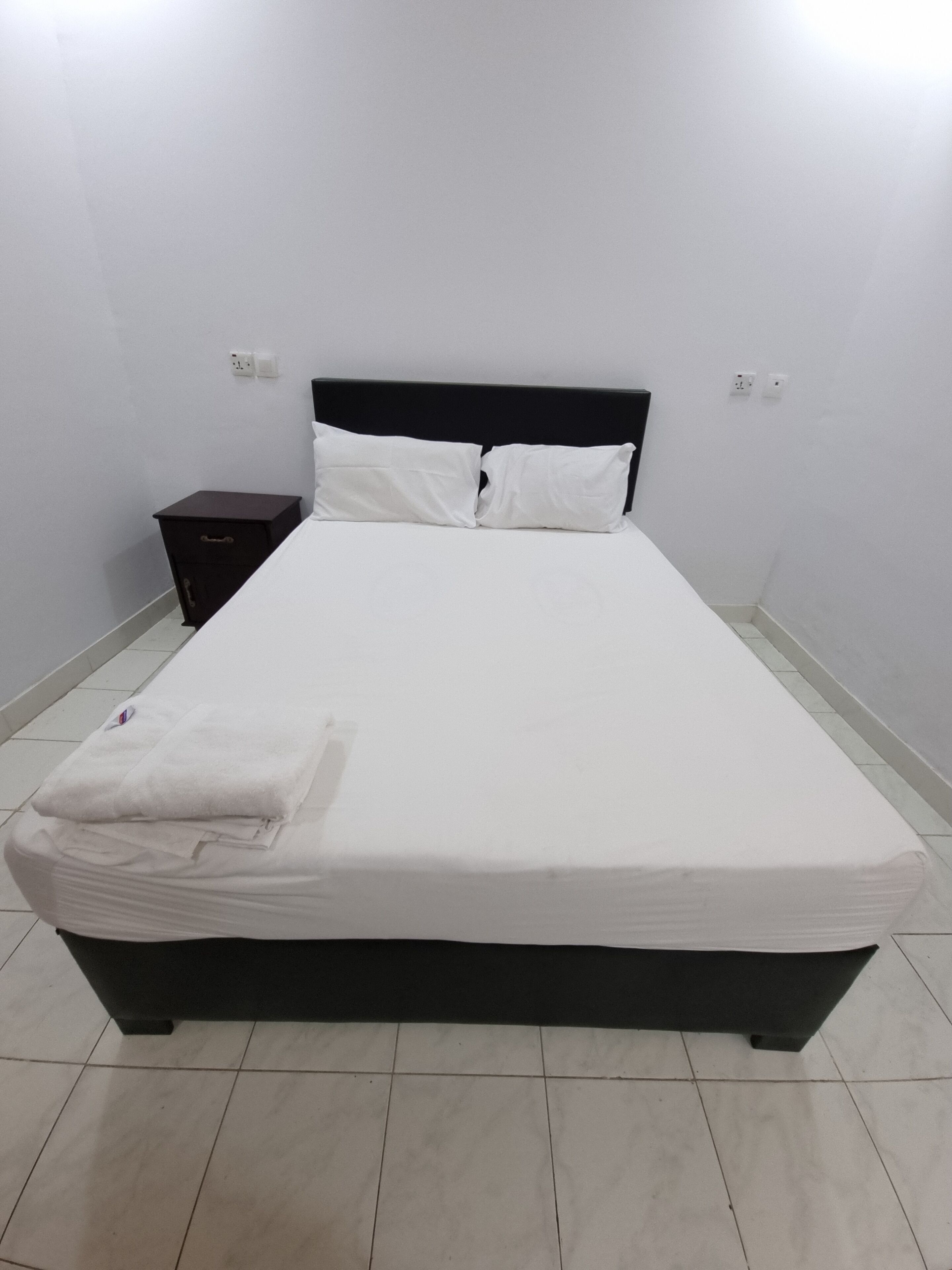 Una cama bien hecha con sábanas y almohadas blancas, un cabecero oscuro y una mesita de noche pequeña con un cajón.
