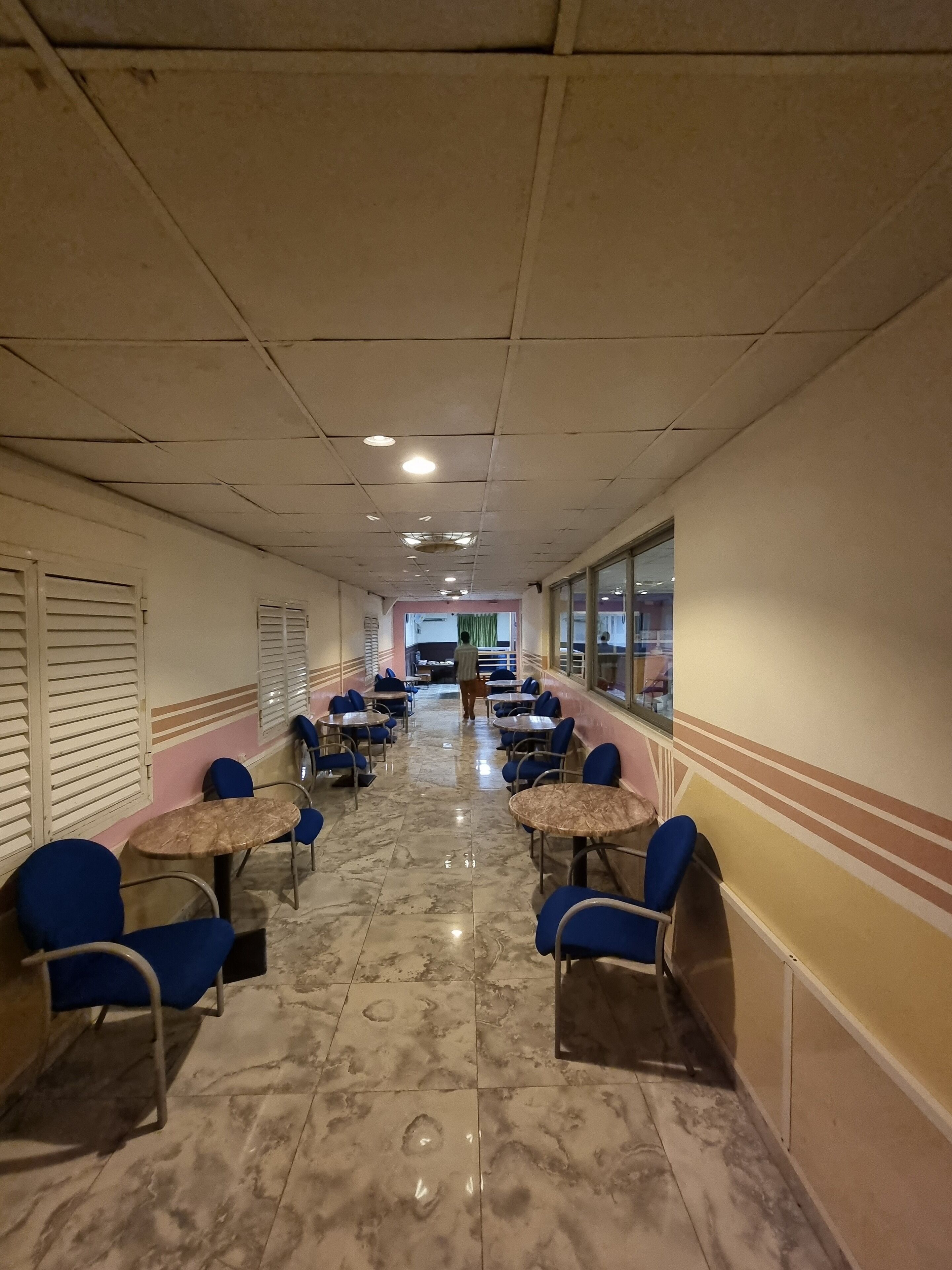 Un pasillo con mesas y sillas, suelo de baldosas e iluminación empotrada en el techo.