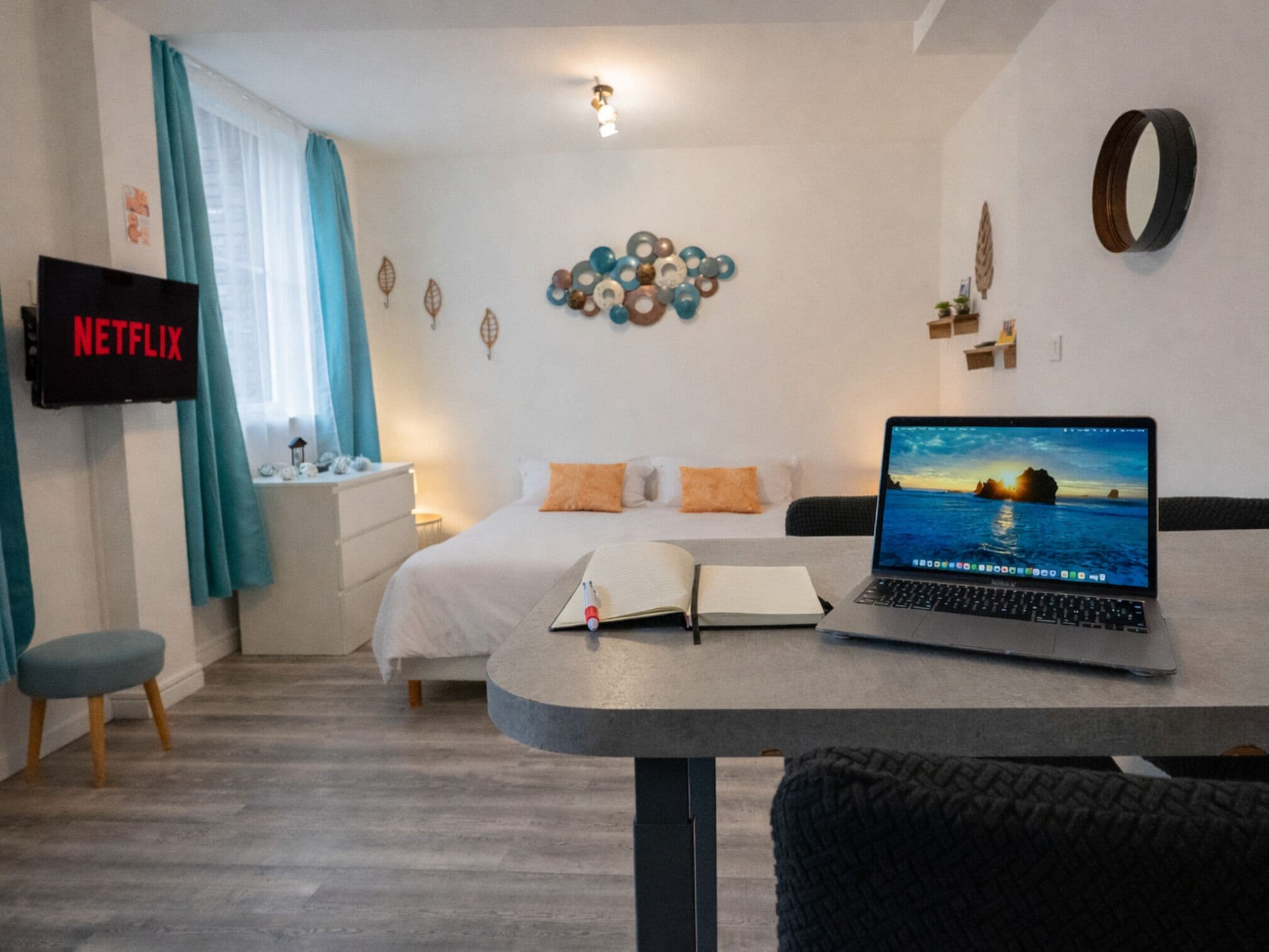 Un dormitorio con una cama, un escritorio con una computadora portátil, una mesita de noche con un libro, un televisor en la pared y una obra de arte decorativa.