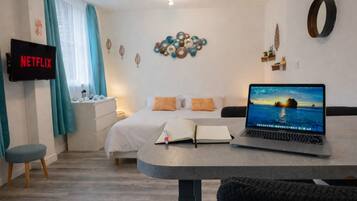 Un dormitorio con una cama, un escritorio con una computadora portátil, una mesita de noche con un libro, un televisor en la pared y una obra de arte decorativa.