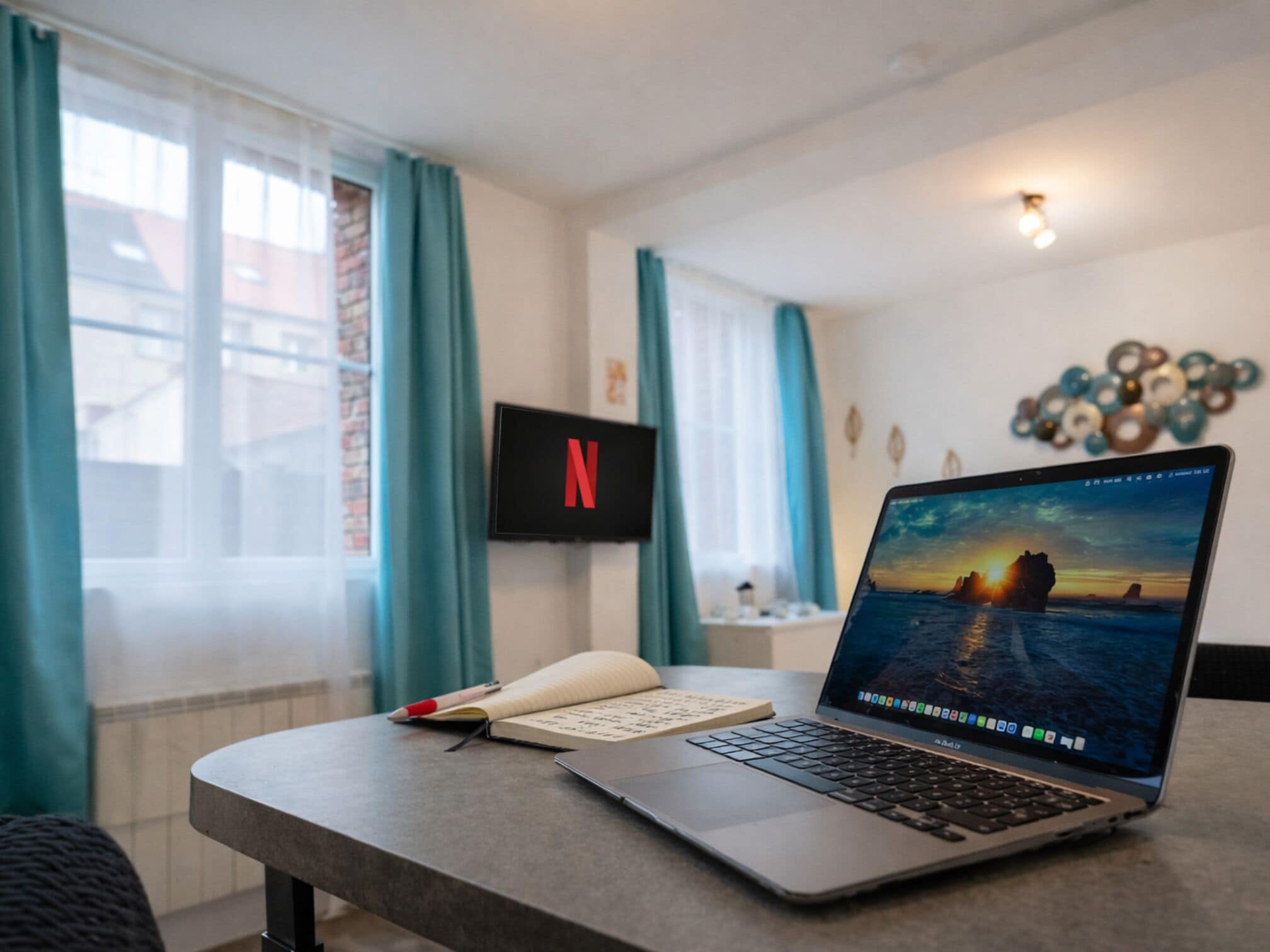 Una computadora portátil sobre una mesa con un cuaderno y un bolígrafo, un televisor con el logotipo de Netflix y un adorno de pared.