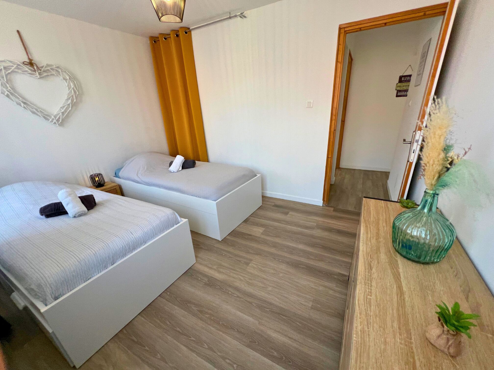 Une pièce avec deux lits, un plancher en bois, une petite table sur laquelle se trouve un vase, et une porte qui donne sur une autre pièce.