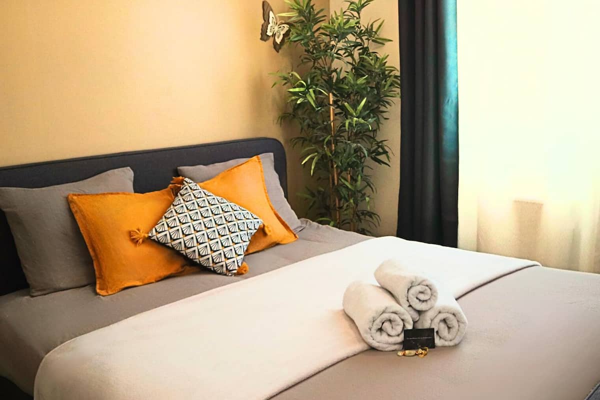 Un lit bien fait, avec un oreiller à motifs gris et blancs, un oreiller de couleur moutarde et des serviettes enroulées posées sur le lit.