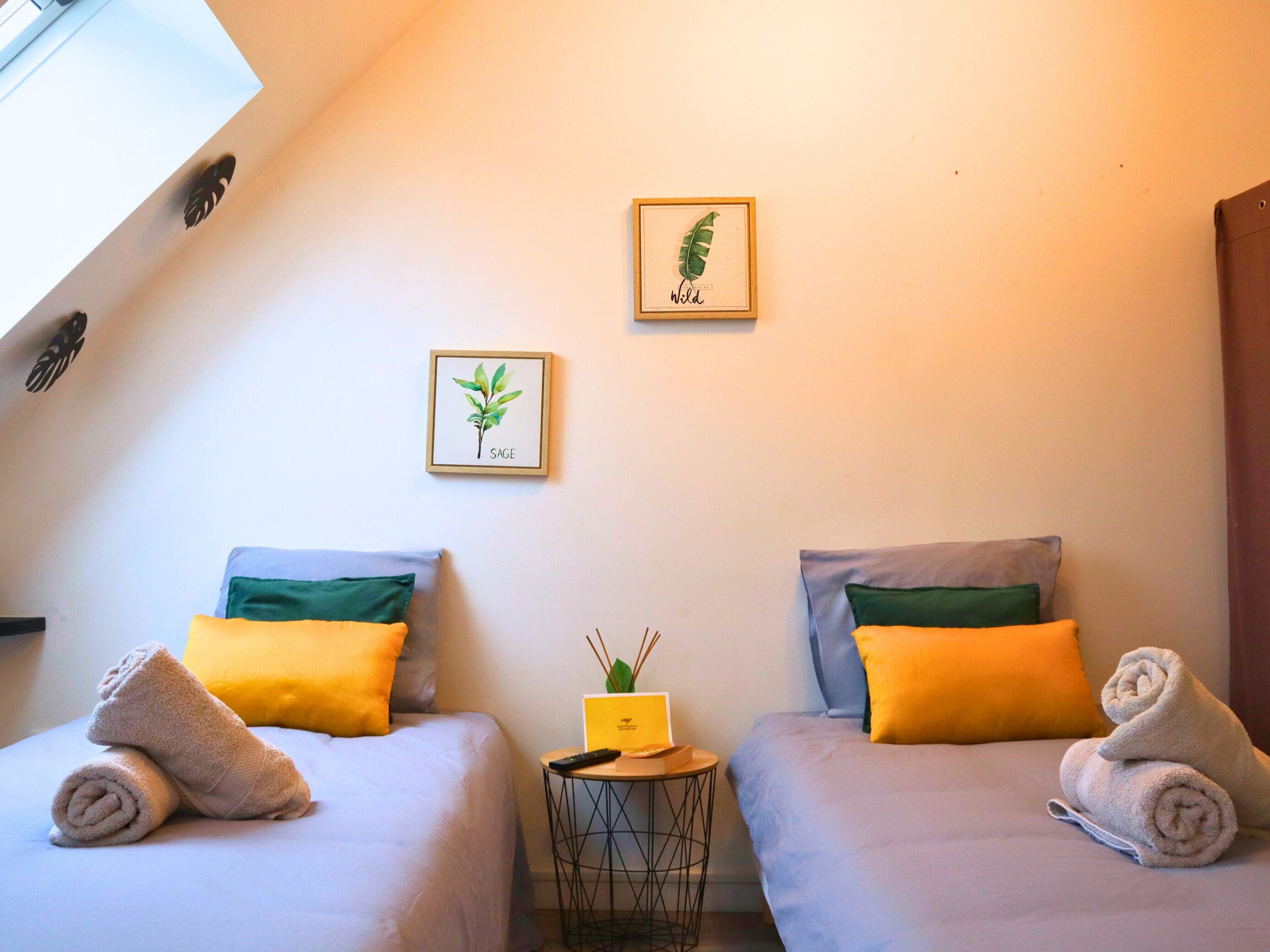 Een kamer met twee bedden, elk voorzien van een opgerolde handdoek, een klein tafeltje met een boek en een plant, en ingelijste botanische prints aan de muur.