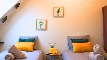 Een kamer met twee bedden, elk voorzien van een opgerolde handdoek, een klein tafeltje met een boek en een plant, en ingelijste botanische prints aan de muur.