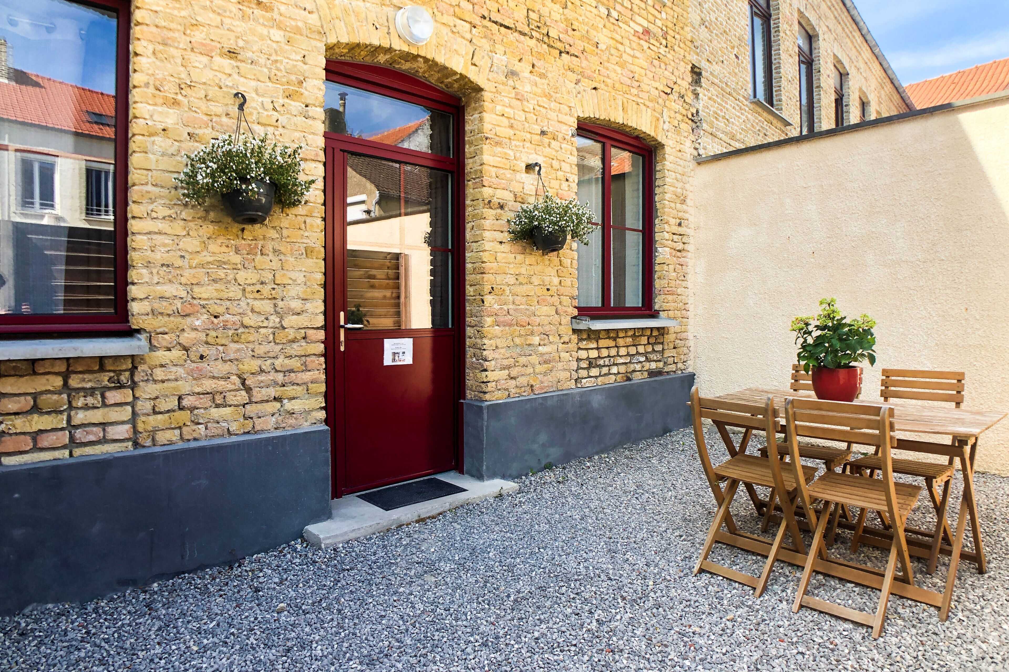 A brick building with a red door, two windows with hanging flower baskets, and a wooden table and chairs outside.