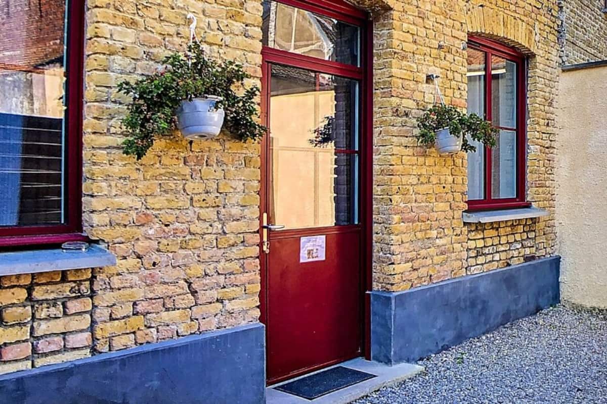 Un bâtiment en briques avec une porte rouge, deux fenêtres ornées de plantes suspendues et un petit espace extérieur aménagé avec des tables et des chaises en bois.