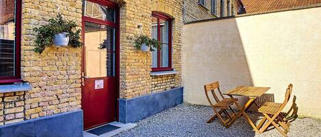 Un edificio de ladrillo con puerta roja, dos ventanas decoradas con plantas colgantes y una pequeña zona exterior con mesas y sillas de madera.