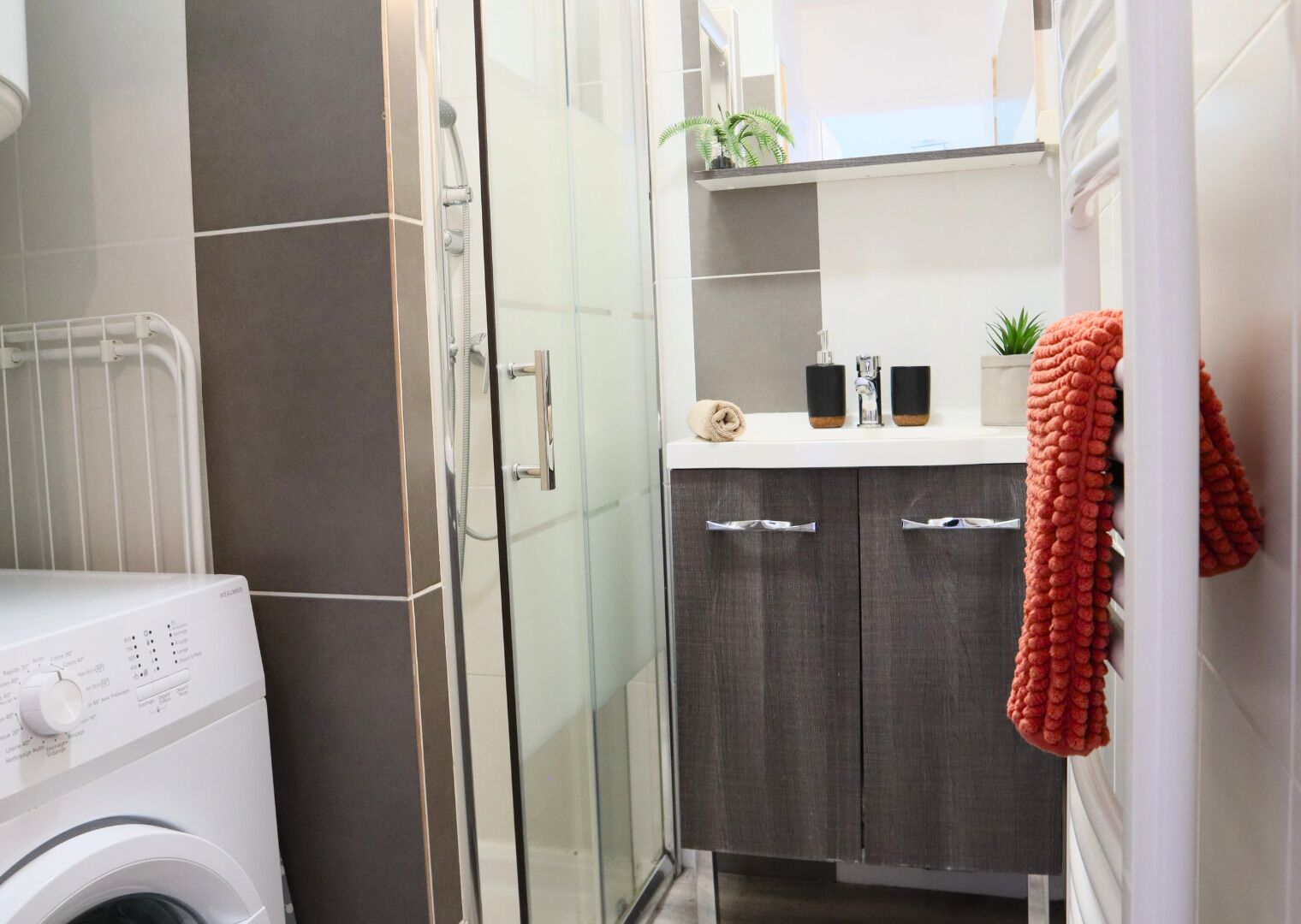 Un baño compacto con cabina de ducha de vidrio, lavadora, armarios de madera oscura y una pequeña encimera con artículos de aseo.