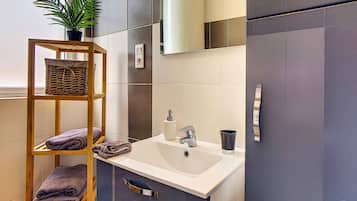 Une salle de bain moderne avec une étagère en bois où sont posées des serviettes, un lavabo avec un miroir au-dessus et une plante en pot.