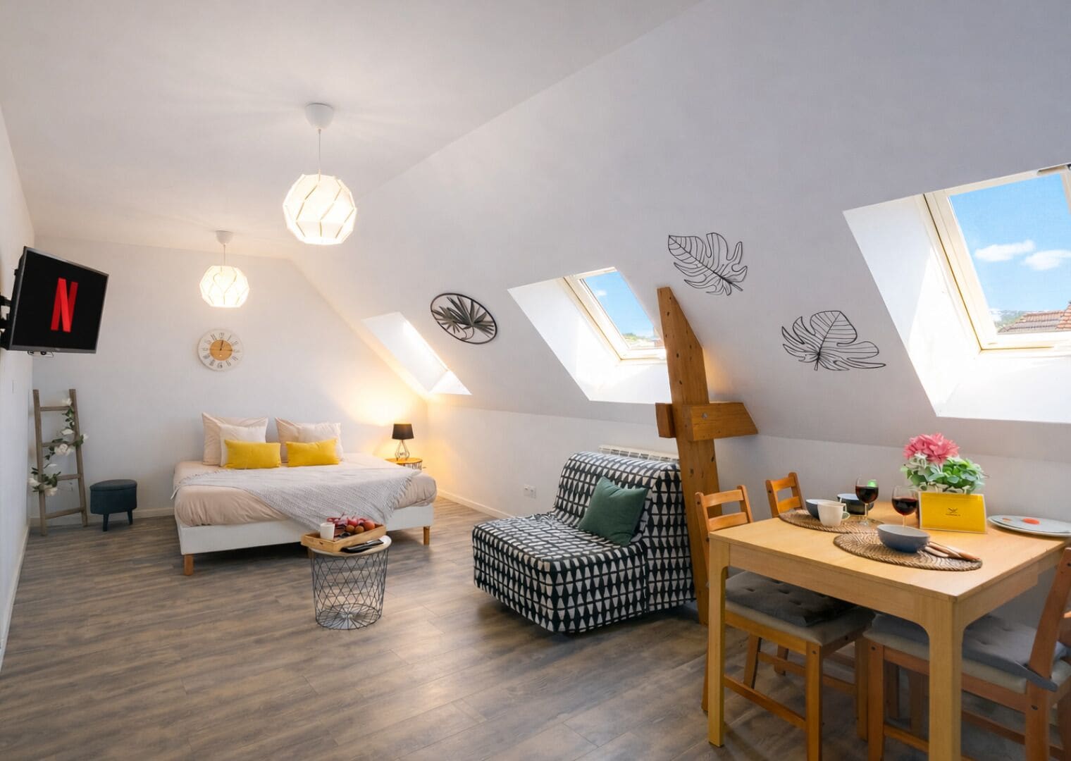A modern loft bedroom with a bed, a desk, and a dining area. There are skylights, a TV, and a clock on the wall.