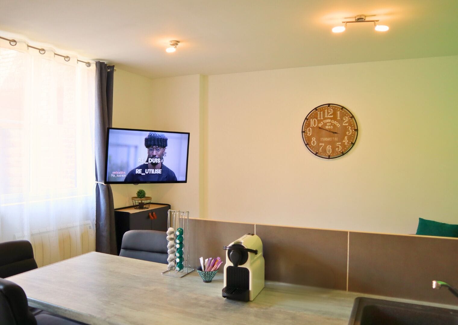 A hotel room with a television, a clock, a table with a speaker, and a plant.