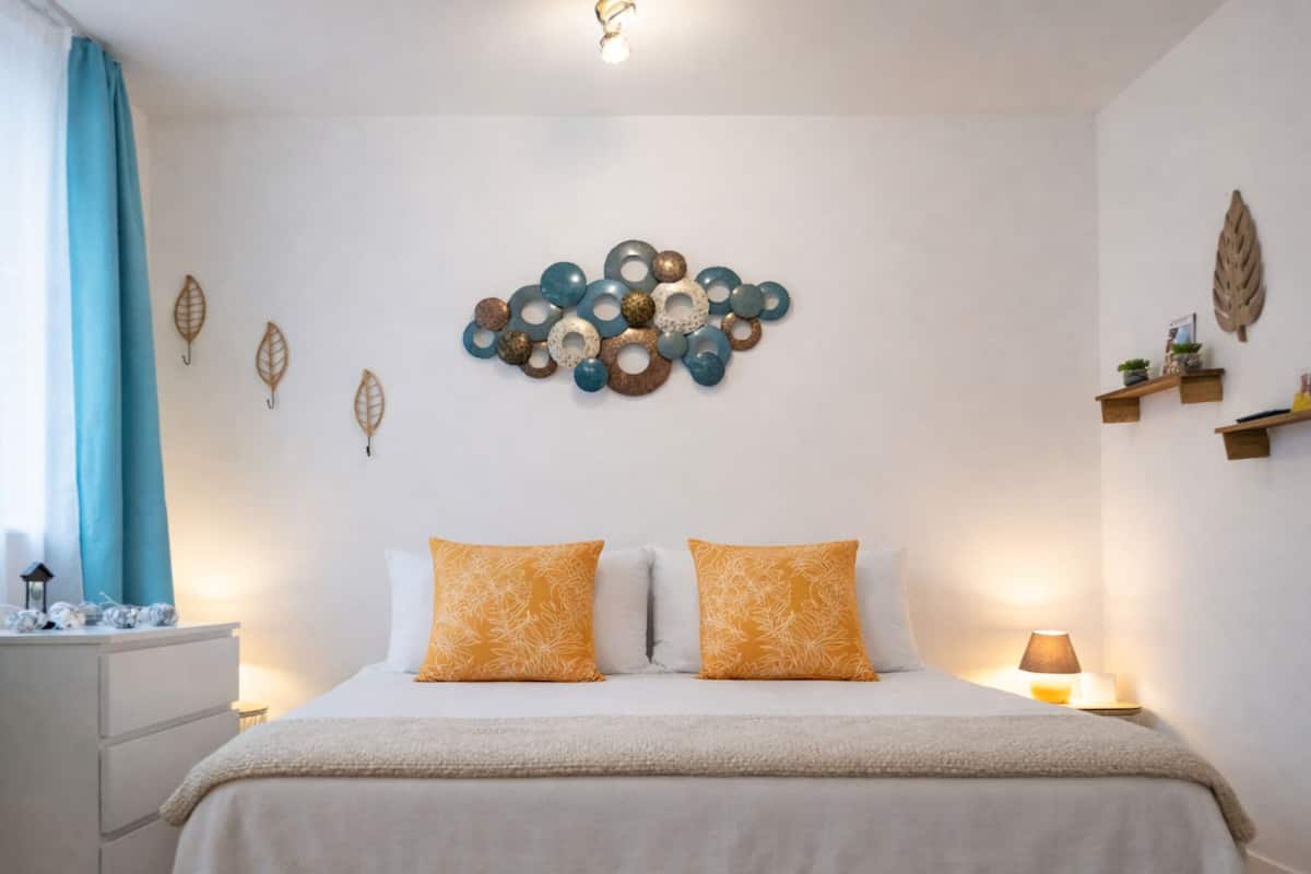 Une chambre à coucher avec un lit, deux oreillers, un élément de décoration murale, une table de chevet, une lampe et une fenêtre avec des rideaux.