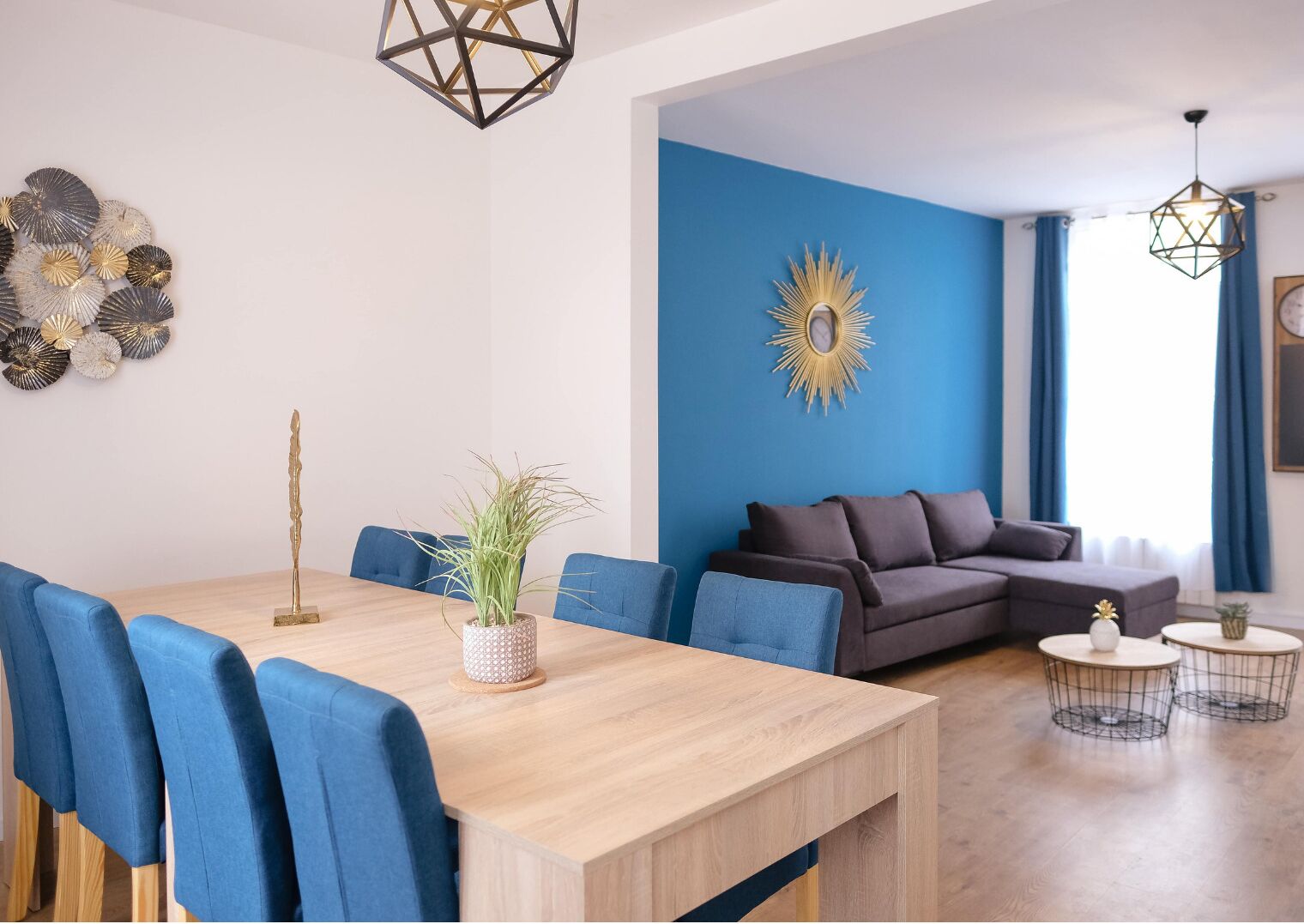 A modern living room with a blue accent wall, a wooden dining table, blue upholstered chairs, a dark sofa, and a decorative sunburst mirror.