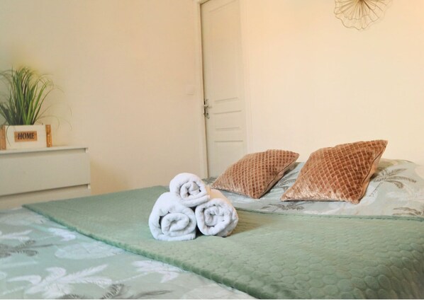 Una cama bien hecha con toallas enrolladas, dos almohadas estampadas y una colcha verde.
