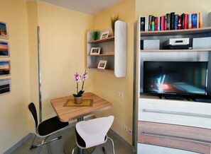 Un salon moderne avec un téléviseur à écran plat, une table à manger en bois, deux chaises, une petite plante en pot et une bibliothèque contenant des livres et des objets décoratifs.