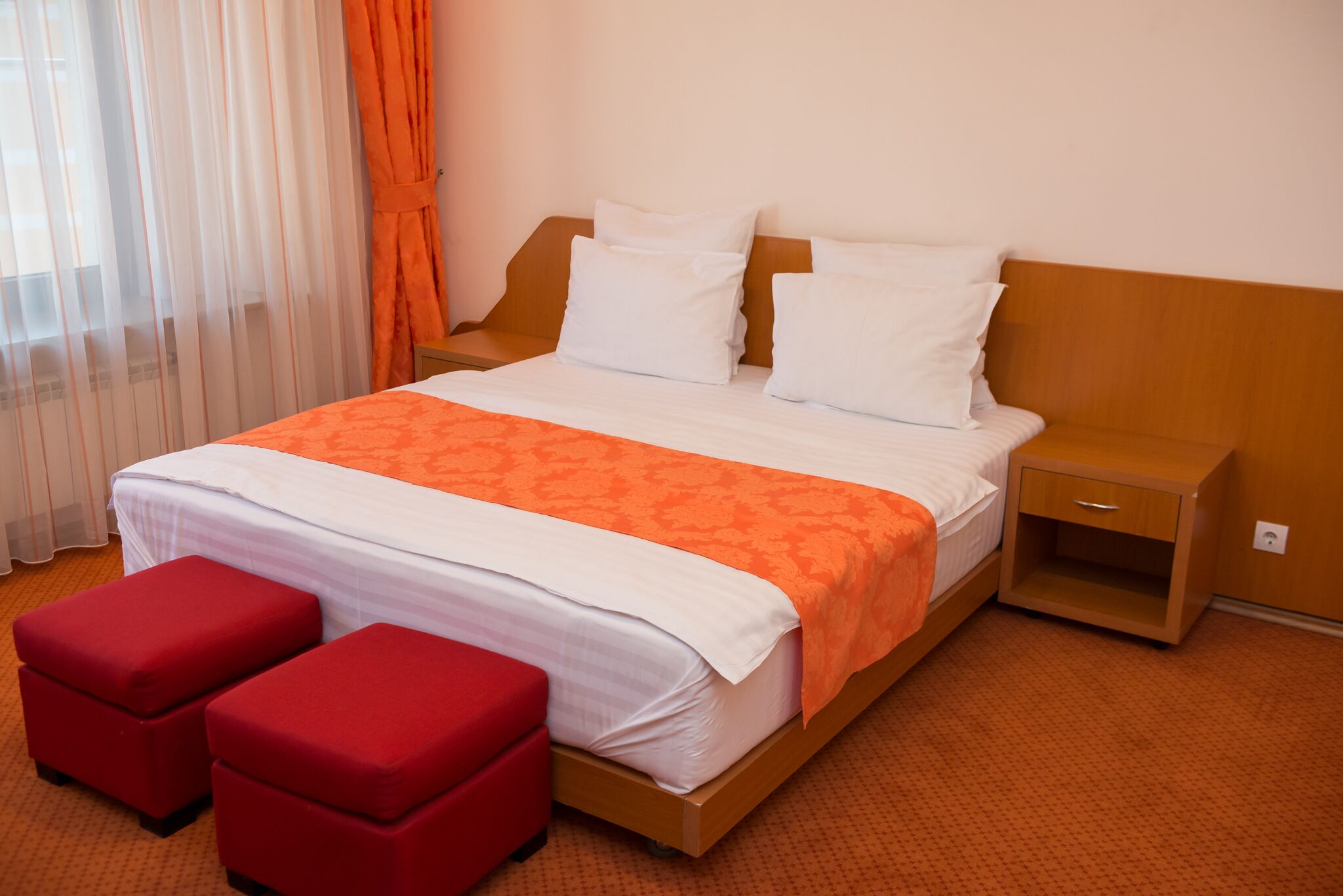 A hotel room with a bed, two red ottomans, a wooden nightstand, and a window with curtains.