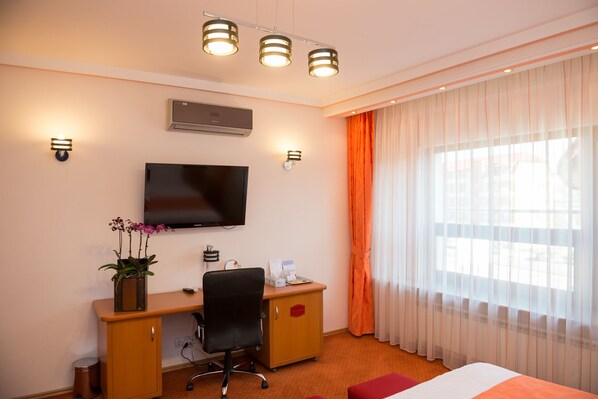 A hotel room with a bed, a desk with a chair, a television, a small cabinet, and a window with curtains.