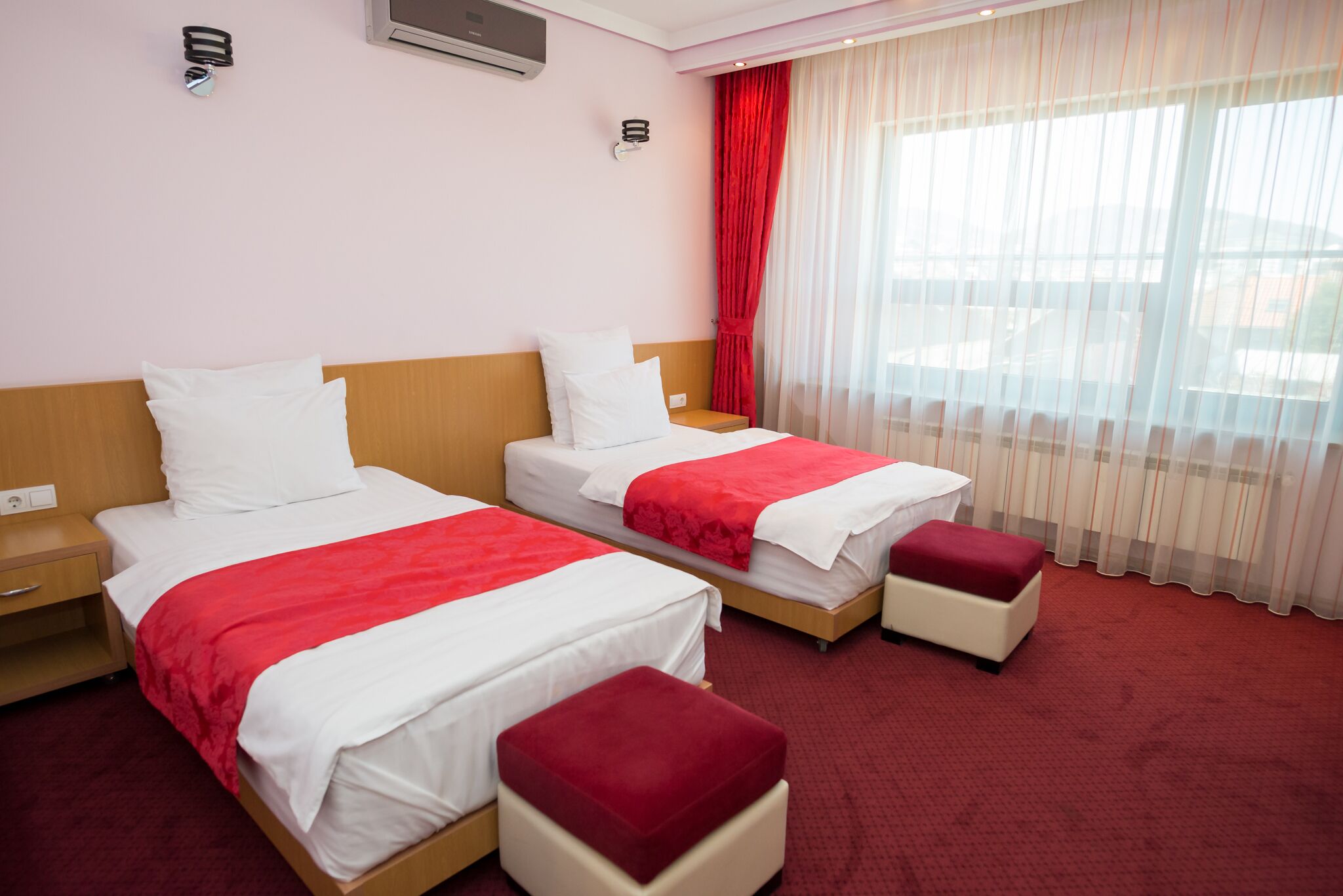 A hotel room with two beds, each with white bedding and red accents, a wooden headboard, a red carpet, and a window with sheer curtains.